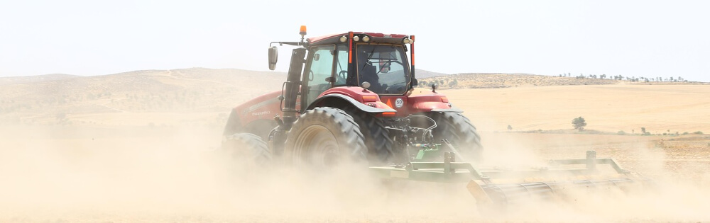Ein roter Traktor fährt durch ein Feld und wirbelt dabei viel Staub auf. Der Hintergrund zeigt eine hügelige, teilweise karge Landschaft unter einem bewölkten Himmel.