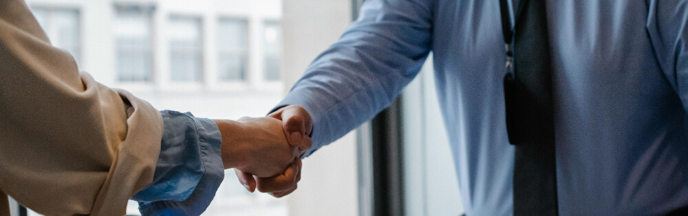 Zwei Personen im formellen Outfit schütteln sich die Hand, im Hintergrund ein Fenster mit verschwommener Aussicht.