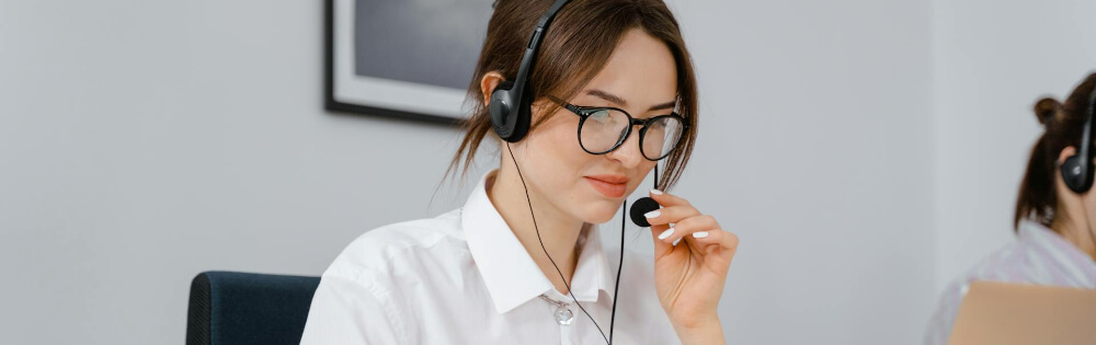 Eine Frau mit Brille und Headset sitzt an einem Schreibtisch und spricht in ein Mikrofon. Im Hintergrund ist eine andere Person zu sehen, die an einem Laptop arbeitet.