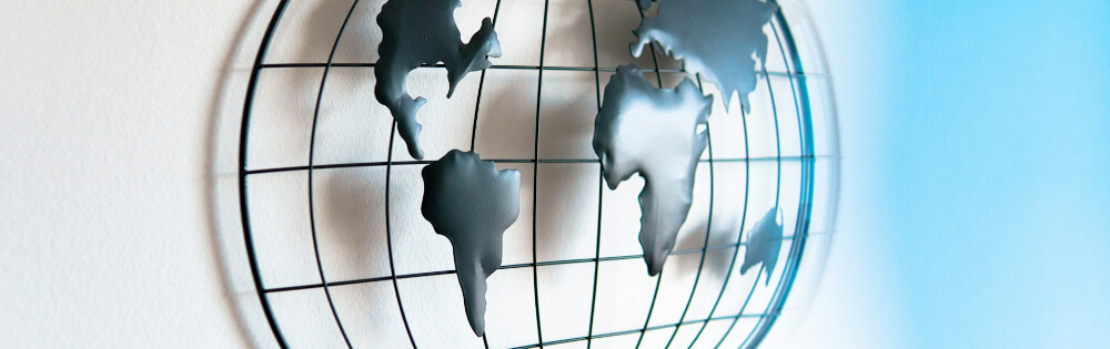 Ein kunstvolles Metallgitter in Form einer Weltkarte hängt an einer weißen Wand. Die Kontinente sind als flache, dunkle Silhouetten dargestellt, die an einem runden Gitterrahmen befestigt sind.